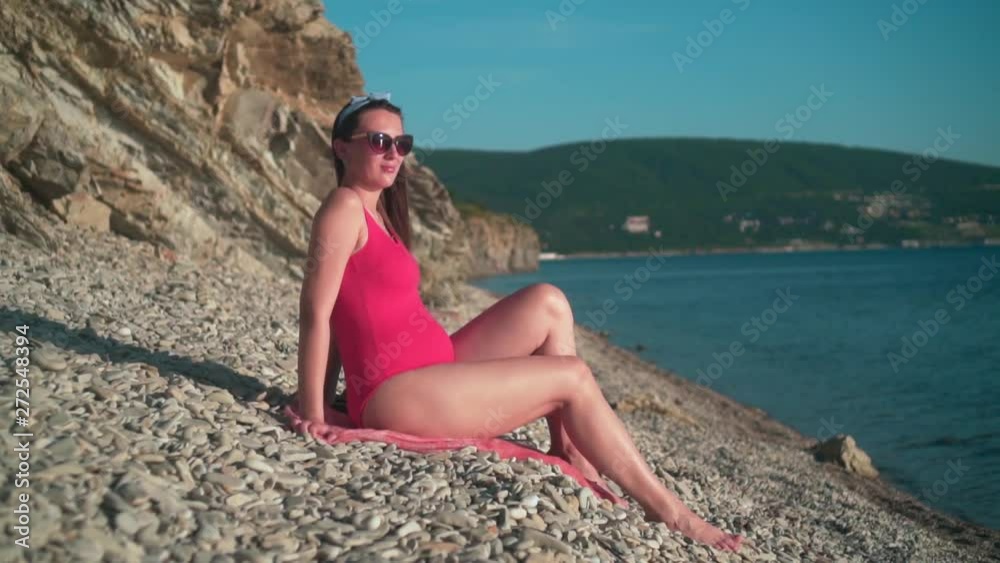 Beautiful pregnant girl in a red one-piece swimsuit and glasses sits on the beach. Mountains in the background. Beautiful pregnant girl in a red one-piece swimsuit and glasses sits on the beach. Mountains in the background.