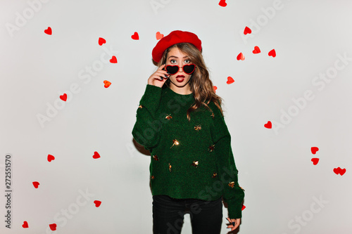 Surprised brunette woman in funny sunglasses isolated on white background decorated with hearts. Indoor photo of emotional girl in red beret and green knitted attire.