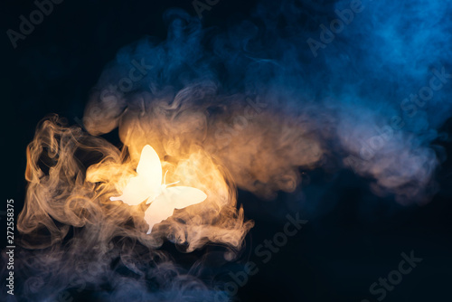 Shining butterfly from paper on a dark background with gradient warm and cold smoke. Magical creature concept with copy space