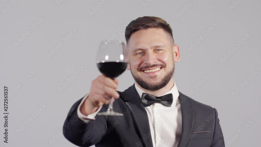 Portrait shot of happy bearded man in suit and bowtie turning towards camera and raising wine glass while posing against grey background Portrait shot of happy bearded man in suit and bowtie turning towards camera and raising wine glass while posing against grey background