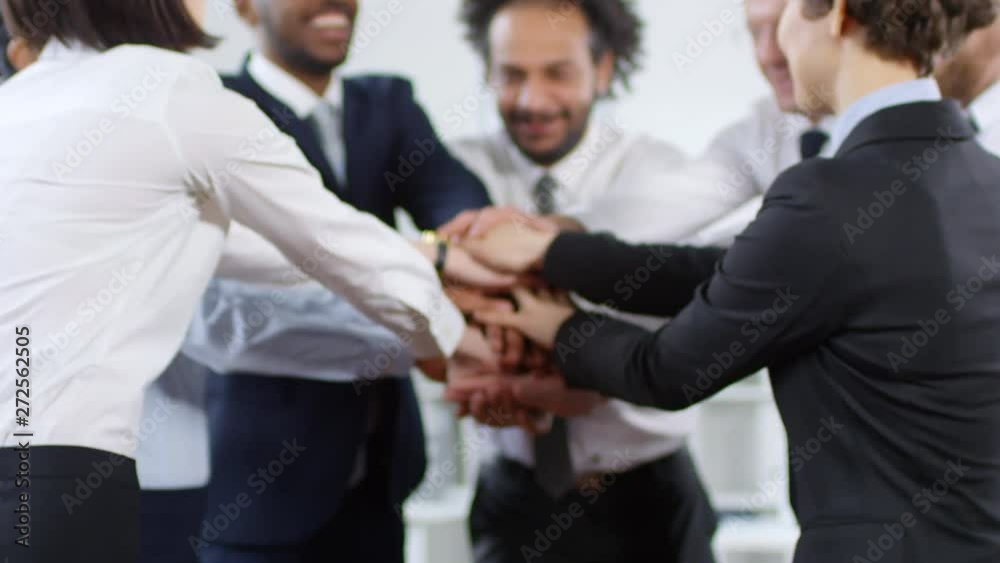 Dolly-in shot of multi-ethnic group of happy businessmen and businesswomen in formalwear standing in office and stacking their hands together, then cheering and smiling Dolly-in shot of multi-ethnic group of happy businessmen and businesswomen in formalwear standing in office and stacking their hands together, then cheering and smiling