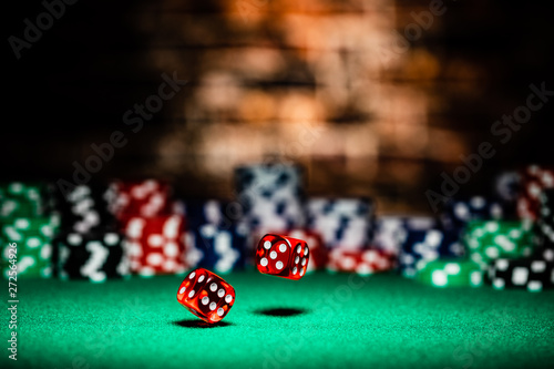 A very close up shot of 2 dice rolling on a green felt table top in very crisp focus, showing 4, 5 and 6 on the faces.  The background is a very soft focused wall of first chips, then a brick wall.