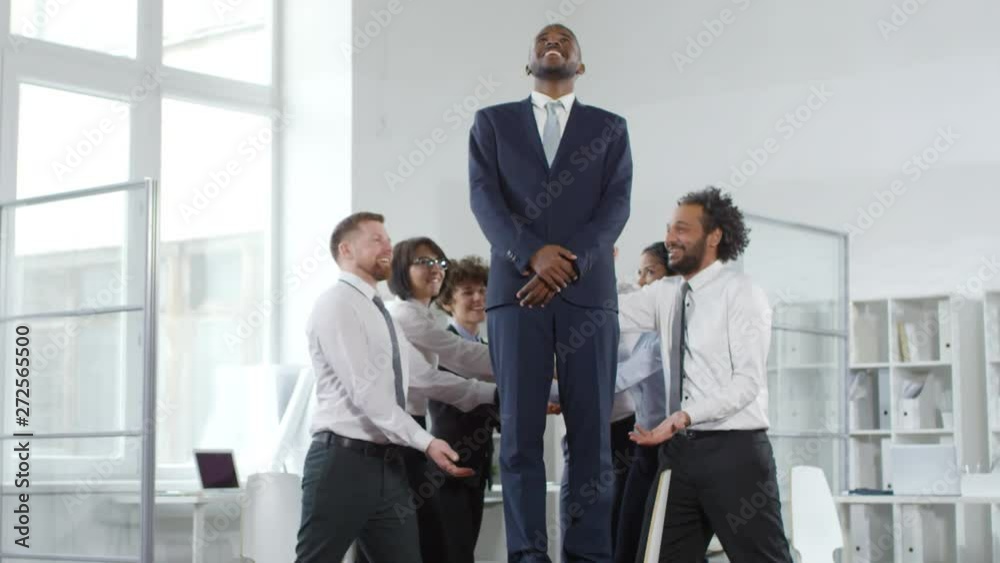 Handheld dolly-in of happy black businessman in suit standing on chair and trust falling into arms of businesspeople. They are catching him and clapping as he is laughing and high-fiving them