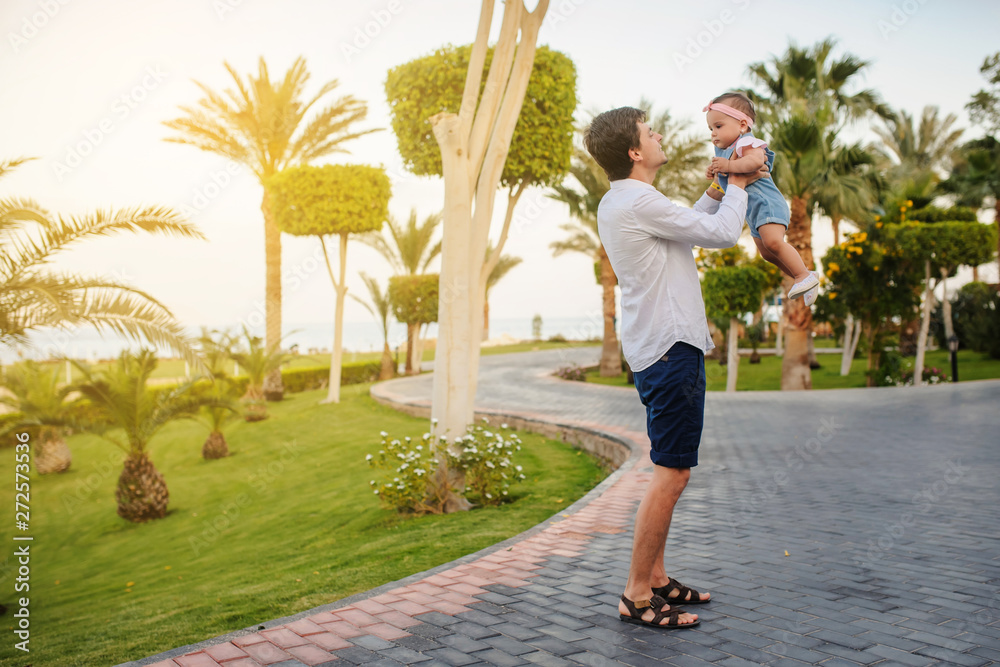 lateral view of a father holding in his arms his little baby girl and walking in the garden of a hotel at a tropic destination