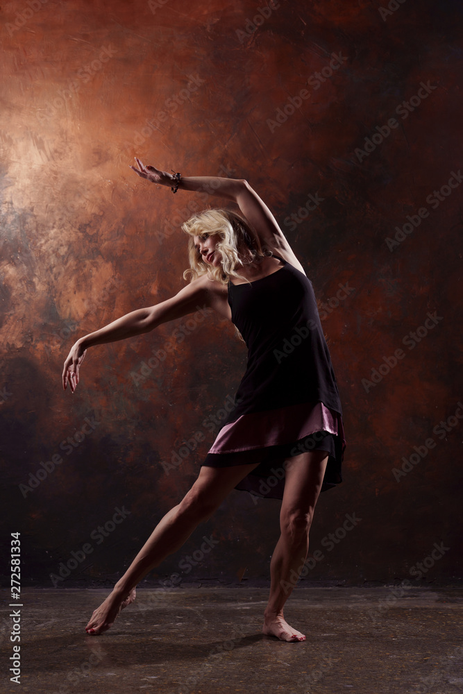 Full-length photo of young dancing blonde in short black dress on brown background