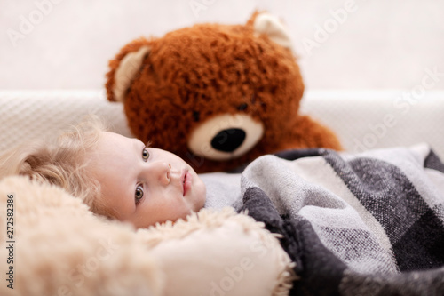 Small baby (a boy of three years old) is lying on the sofa under the rug with a teddy bear toys.