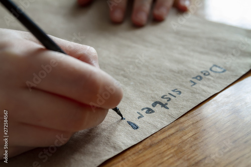 A small girl writes a letter to dear sister by black paint on brown paper. Closeup, selective focus
