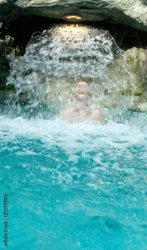 young sexy blond woman in pink bikini enjoys the falling water behind the waterfall in the spa pool