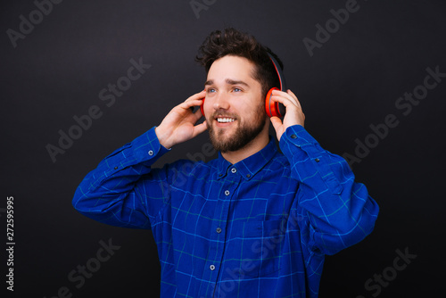 Young bearded man looking aside an listening to the music through a red headset on black background.