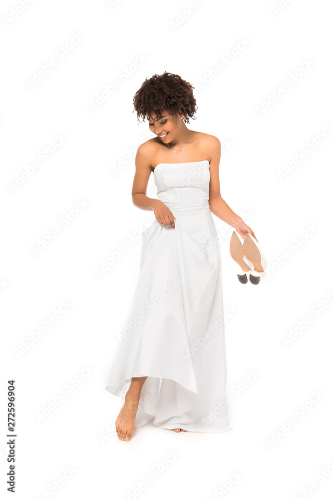 cheerful african american bride touching wedding dress and holding shoes while standing isolated on white cheerful african american bride touching wedding dress and holding shoes while standing isolated on white