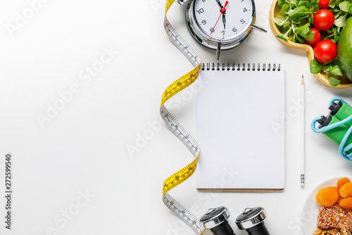 top view of sport equipment  measuring tape  alarm clock and diet food near empty notebook on white background