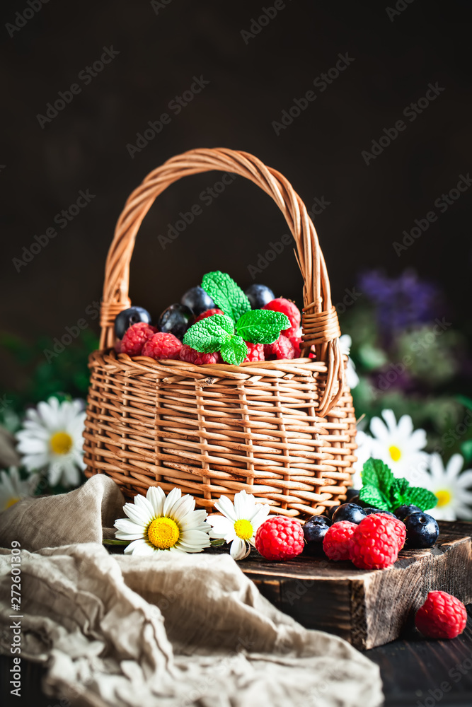 Raspberries and blueberries in a basket with chamomile and leaves on a dark background. Summer and healthy food concept. Selective focus. Background with copy space.