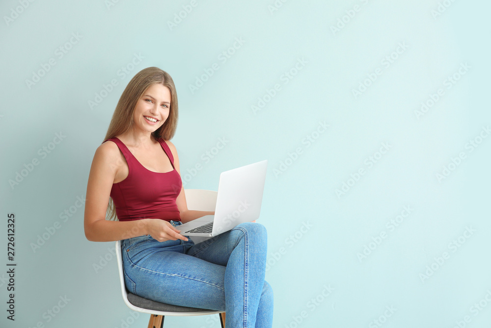 Beautiful young woman with laptop sitting on chair against color background