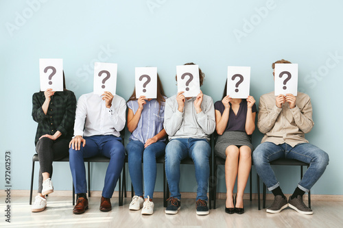 Young people hiding faces behind paper sheets with question marks while waiting for job interview indoors