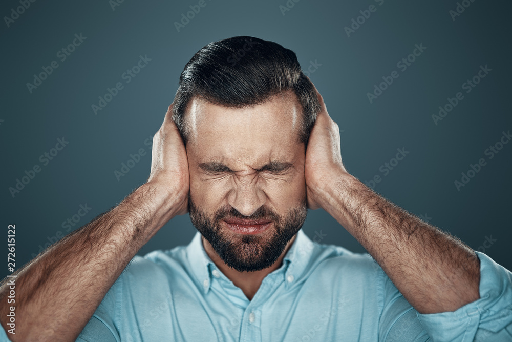 Problems. Frustrated young man covering ears with hands while standing against grey background Problems. Frustrated young man covering ears with hands while standing against grey background