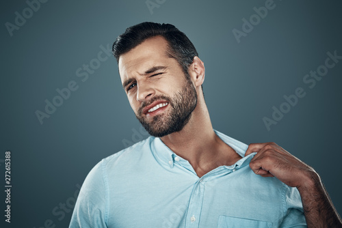 Uncomfortable. Tired young man pulling collar while standing against grey background