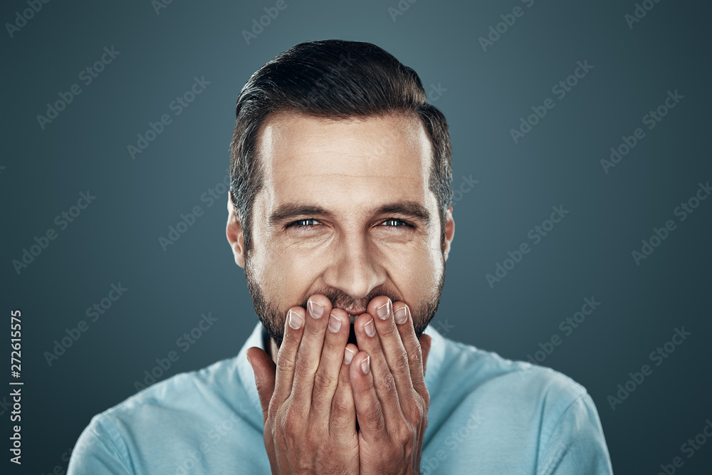 Sincere fun. Handsome young man laughing and covering mouth with hands while standing against grey background Sincere fun. Handsome young man laughing and covering mouth with hands while standing against grey background