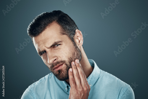 Need to shave. Handsome young man touching his beard and looking at camera while standing against grey background