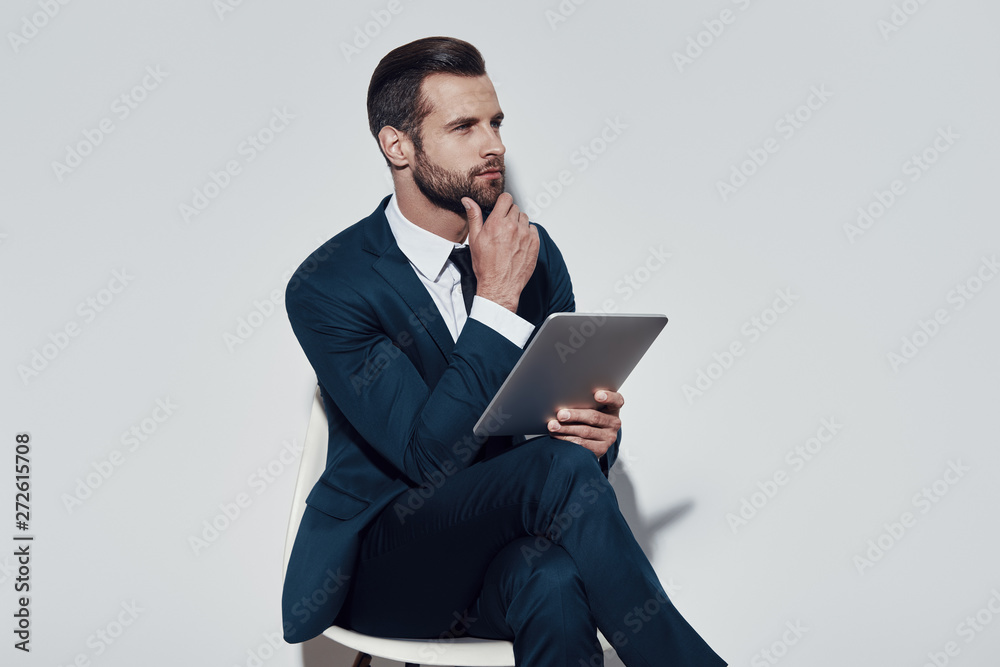 Organizing everything. Handsome young man working using digital tablet and keeping hand on chin while sitting against grey background Organizing everything. Handsome young man working using digital tablet and keeping hand on chin while sitting against grey background