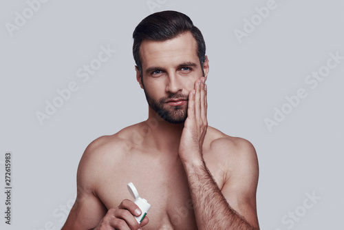 Another beauty treatment. Handsome young man applying moisturizer and looking at camera while standing against grey background