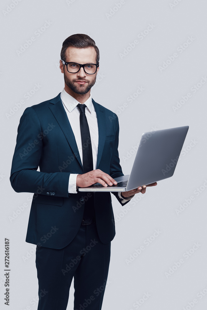 Using technologies. Handsome young man looking at camera and smiling while standing against grey background Using technologies. Handsome young man looking at camera and smiling while standing against grey background