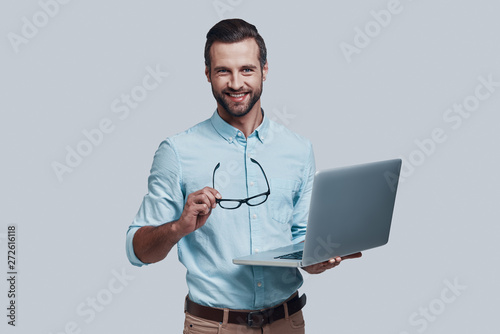 Always ready to help. Good looking young man carrying laptop and looking at camera while standing against grey background