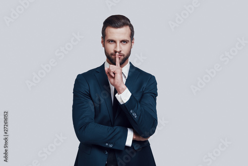 Keep silence! Handsome young man working keeping finger on lips while standing against grey background