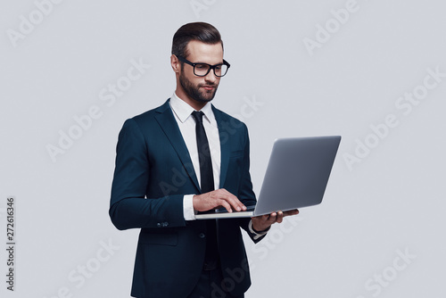 Paying attention to every detail. Handsome young man using laptop and smiling while standing against grey background
