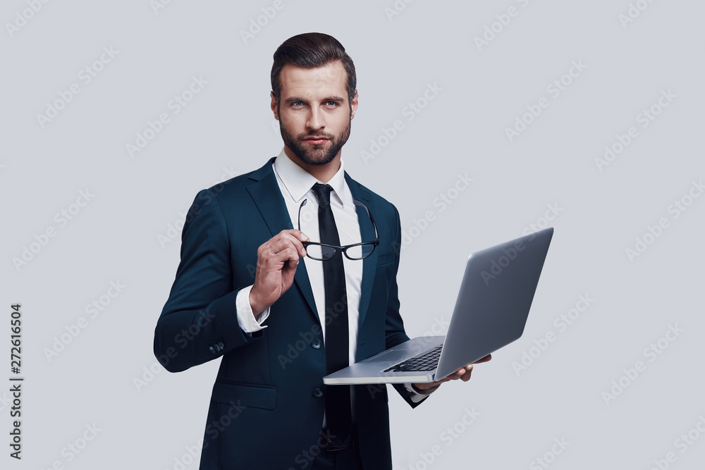 Young business expert. Handsome young man looking at camera and working using laptop while standing against grey background Young business expert. Handsome young man looking at camera and working using laptop while standing against grey background