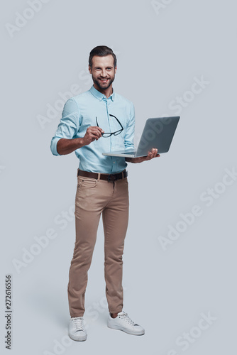 Business is his life. Full length of good looking young man carrying laptop and looking at camera while standing against grey background