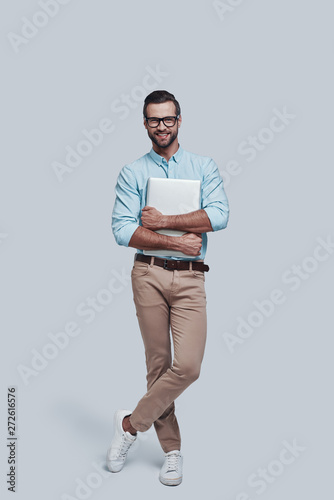 Young and successful. Full length of good looking young man carrying laptop and looking at camera while standing against grey background