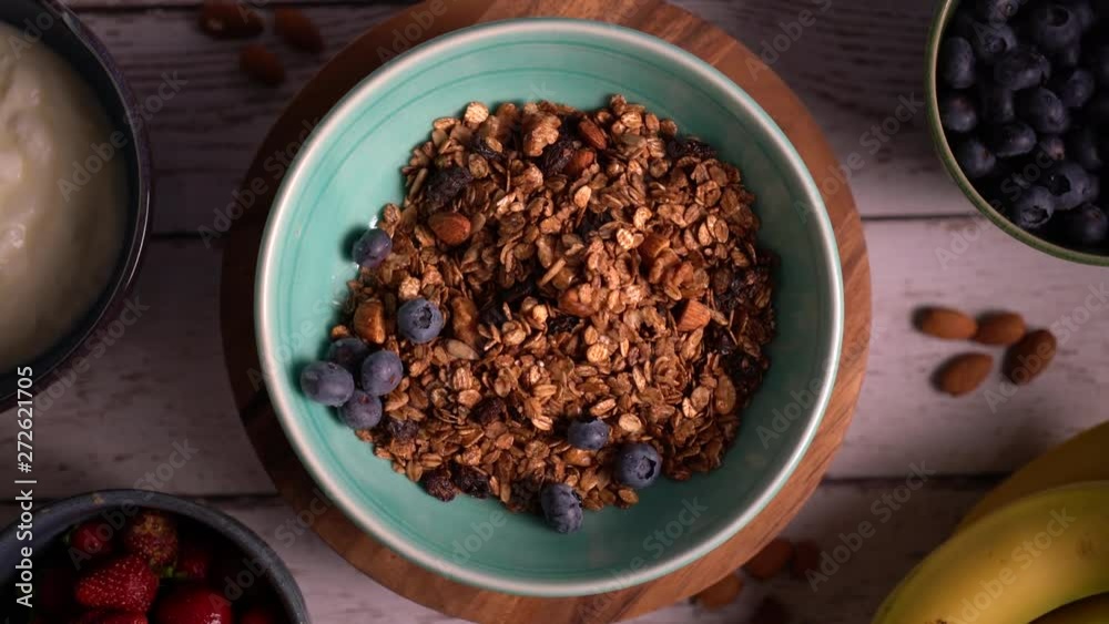 Flat lay of granola turning as hand with spoon puts blueberries in the granola. Flat lay of granola turning as hand with spoon puts blueberries in the granola.