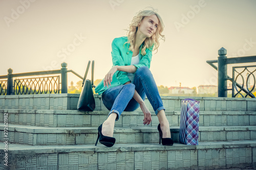 Blonde in blue jeans, turquoise jacket and black high-heeled shoes sits on the steps outside and looks at the sky, next to her are a handbag and shopping bags