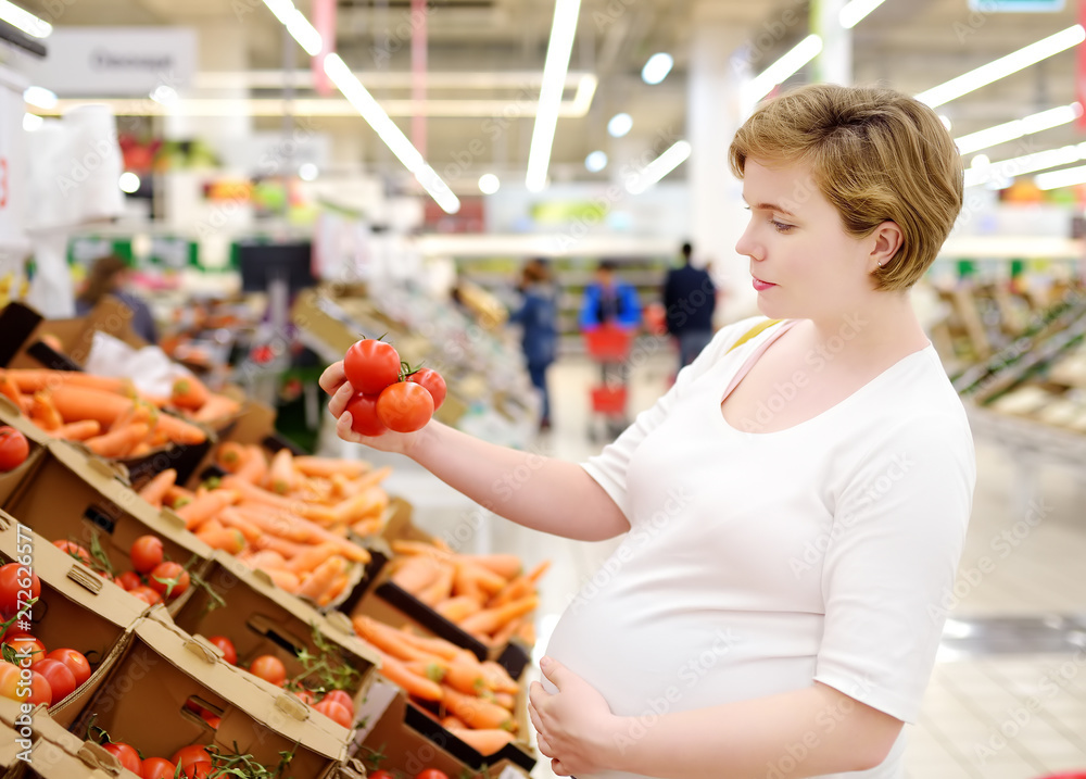Young pregnant woman choosing fresh tomato in supermarket