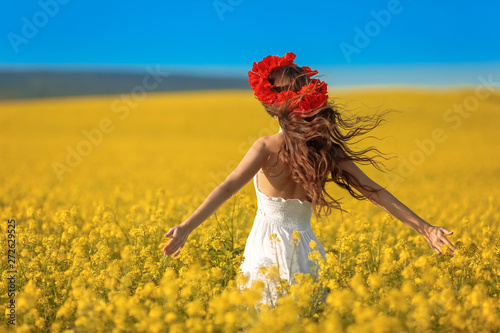 Beautiful young woman with long healthy hair over Yellow rape field landscape background. Attracive brunette girl with red poppy wreath on hairstyle, outdoor portrait. Natural beauty.