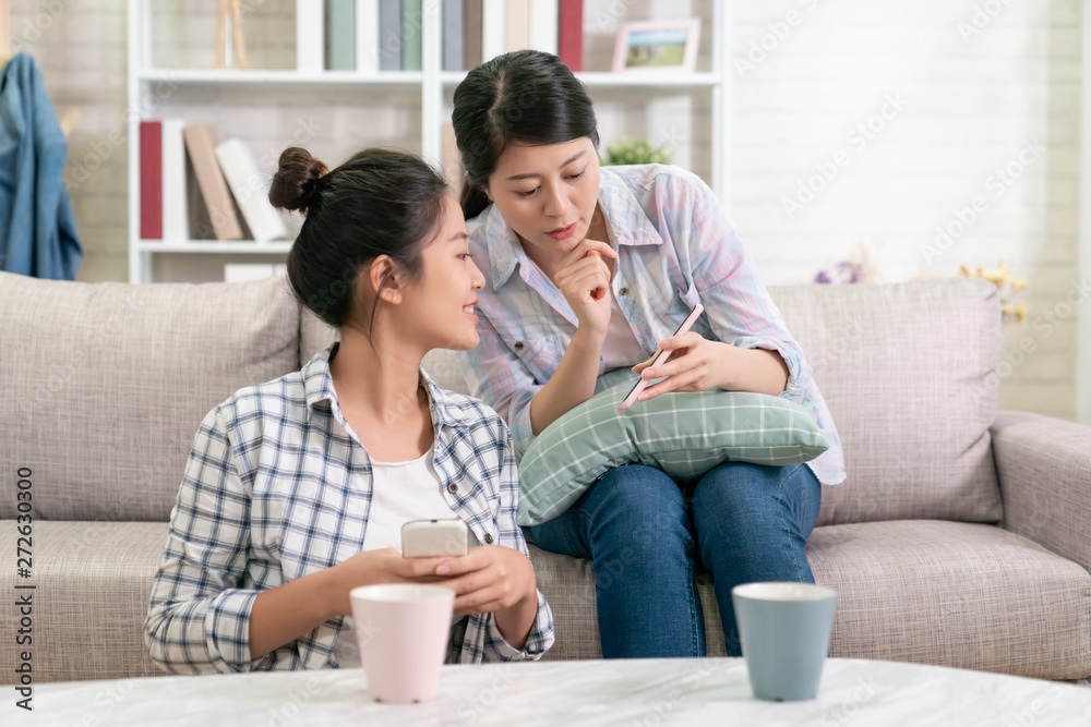 Obraz premium Young asian korean elegant woman showing mobile phone to female friend while sitting on sofa and floor at home in cozy modern living room. two girls relax in apartment on weekends looking cellphone