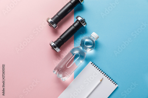 top view of bottle with water, dumbbells and blank notebook on blue and pink background