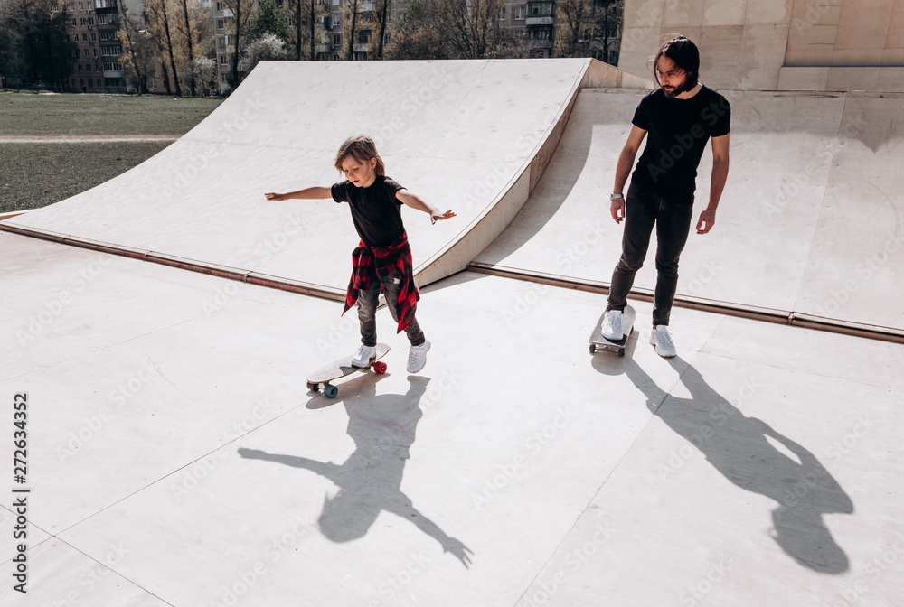 Obraz premium Szczęśliwy ojciec i jego synek ubrani w zwykłe ubrania jeżdżą w słoneczny dzień na deskorolkach w skateparku ze zjeżdżalniami