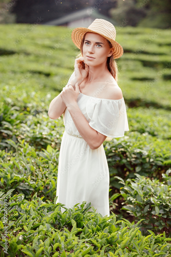 Portrait of a beautiful and romantic young blonde girl in white summer dress and hat posing while walking among green bushes in park during vacation on warm summer day. Concept of love for the natural
