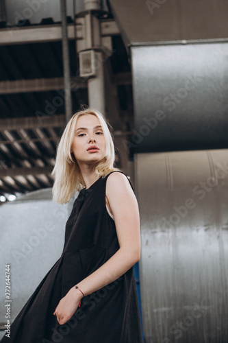 Stylish young girl dressed in a comfortable, daily black dress and white sneakers pose for a photo on an industrial background, urban clothing style. Street photography