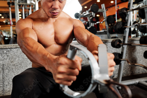 Close-up of young shirtless muscular man sitting on bench and training with weights in fitness club