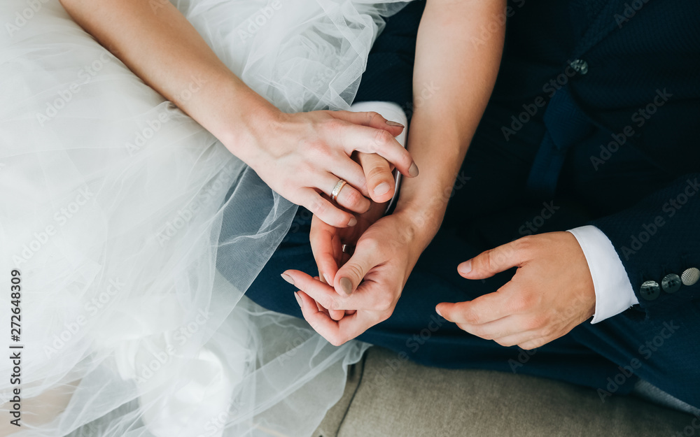 Fototapeta premium Wedding couple sitting on the sofa barefoot. Bride and groom holding hands together. Love story. Elegant clothes. Cosy home interior.