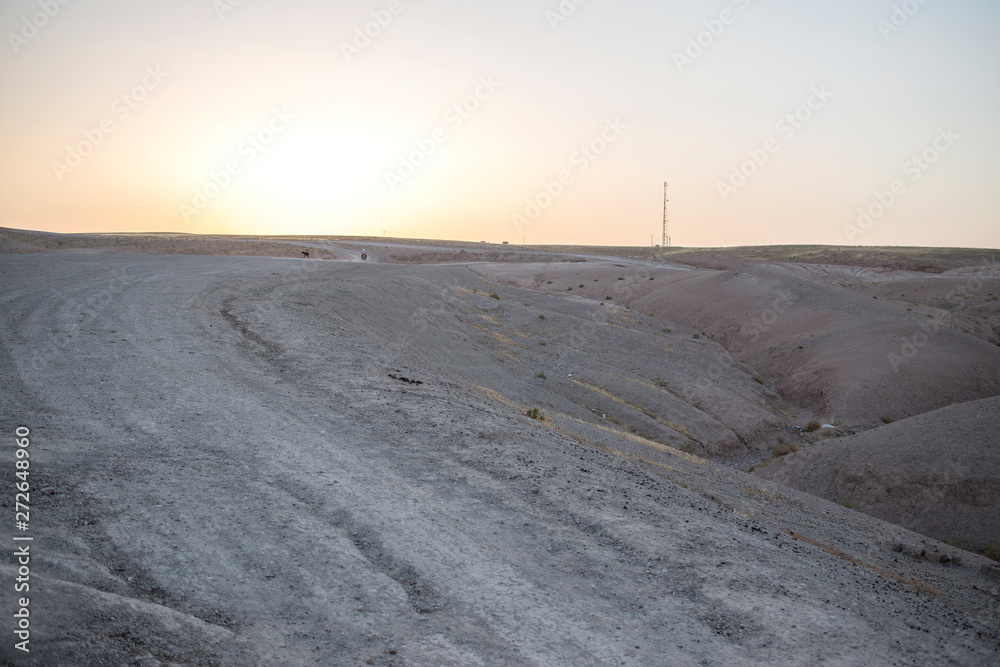 View of a man riding a motorcycle on a dirt road in the Sahara desert in Morocco, Africa. The sun sets behind the mountains.