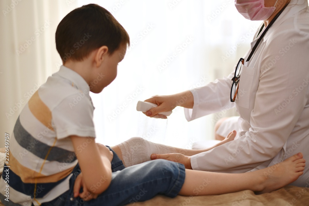 © uly.u.v - A doctor in a white robe bandages the knee of a child sitting in front of her. The boy, waiting, folded his arms and watches the scene. © uly.u.v - A doctor in a white robe bandages the knee of a child sitting in front of her. The boy, waiting, folded his arms and watches the scene.