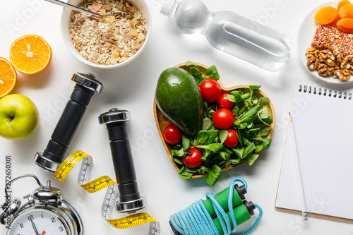 top view of sport equipment, measuring tape, alarm clock and notebook near diet food and water on white background