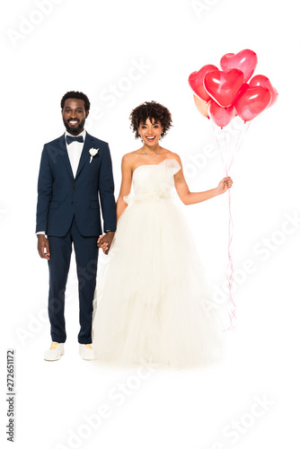 cheerful african american bridegroom holding hands with happy bride with balloons isolated on white