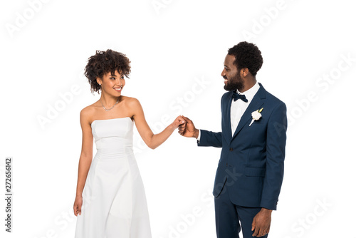 happy african american bridegroom looking at bride while holding hands isolated on white