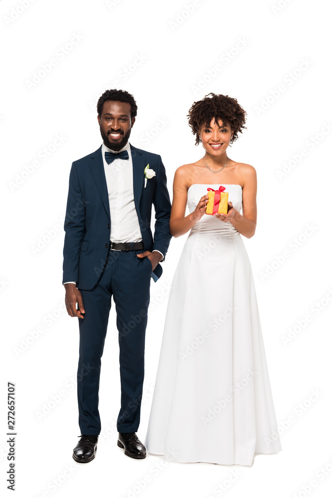 happy african american bridegroom standing with hand in pocket near cheerful bride holding gift box isolated on white
