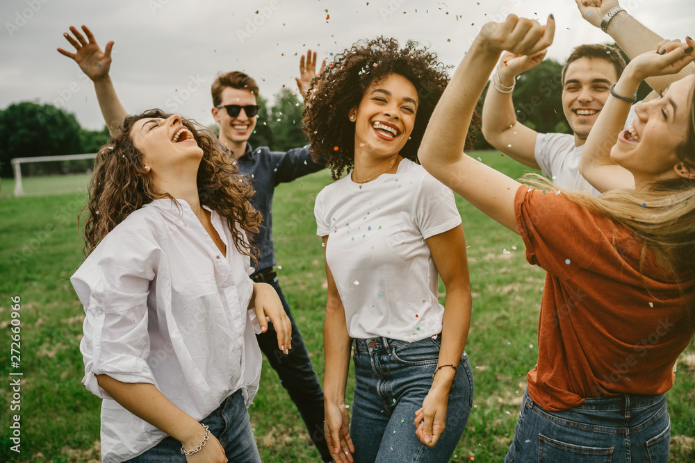 Fototapeta premium Grupa pięciu przyjaciół bawi się w parku - Milenialsi tańczą na łące wśród podrzuconych w powietrze konfetti - Dzień wolności i beztroski
