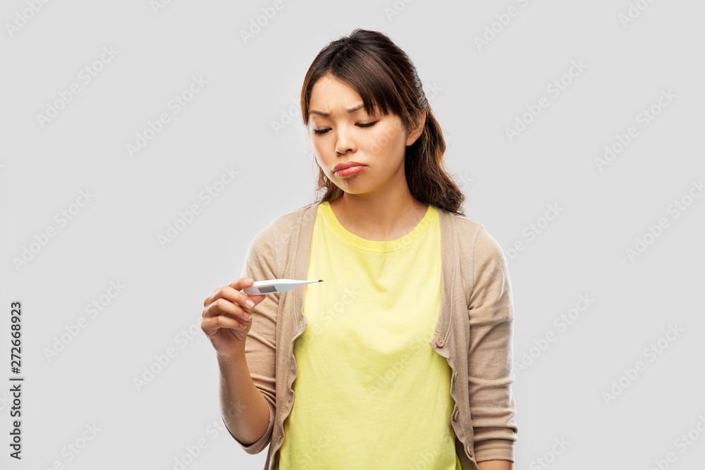health and fever concept - sick asian young woman looking at thermometer over grey background